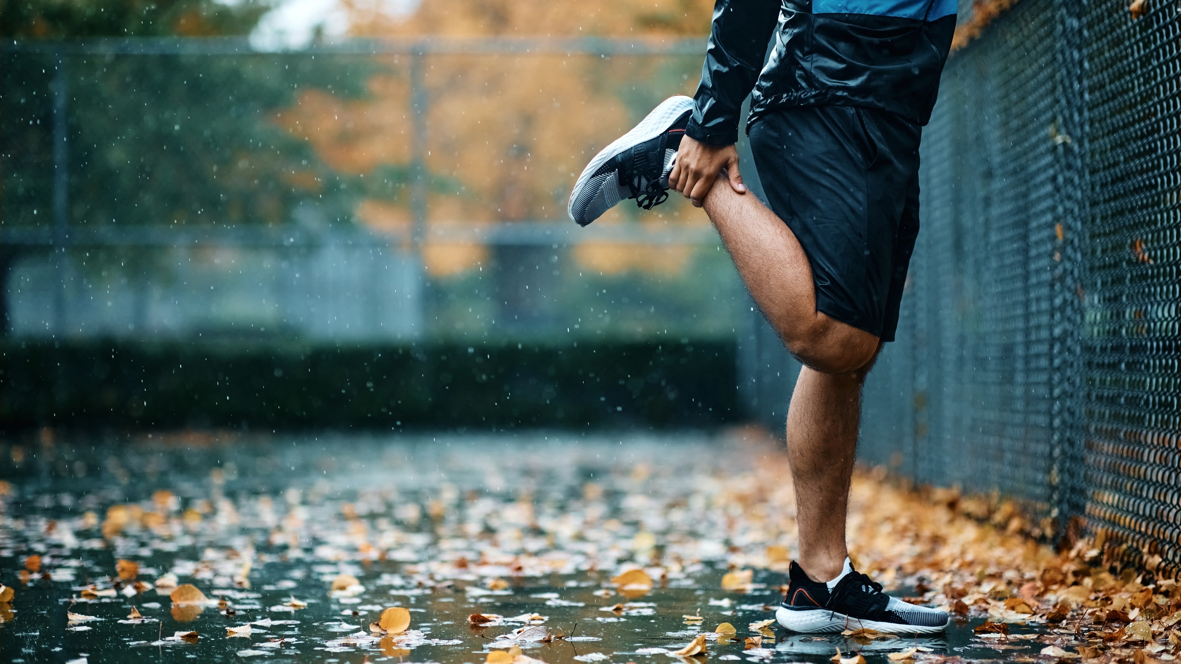 Lower section of a man stretching his quad in a tennis court on a rainy fall day