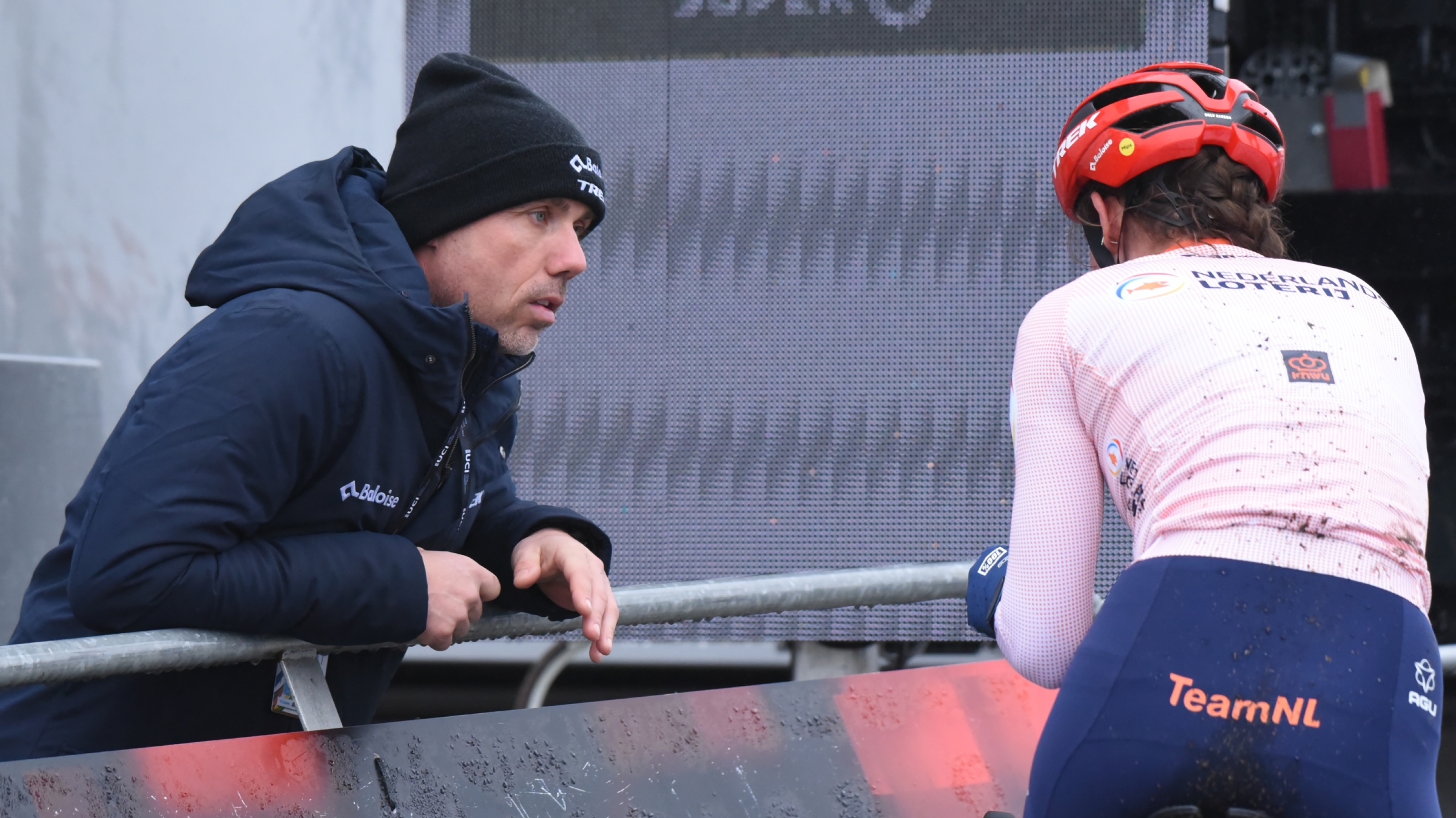 Coach leaning over a railing to speak with a muddy cyclist on the bike.